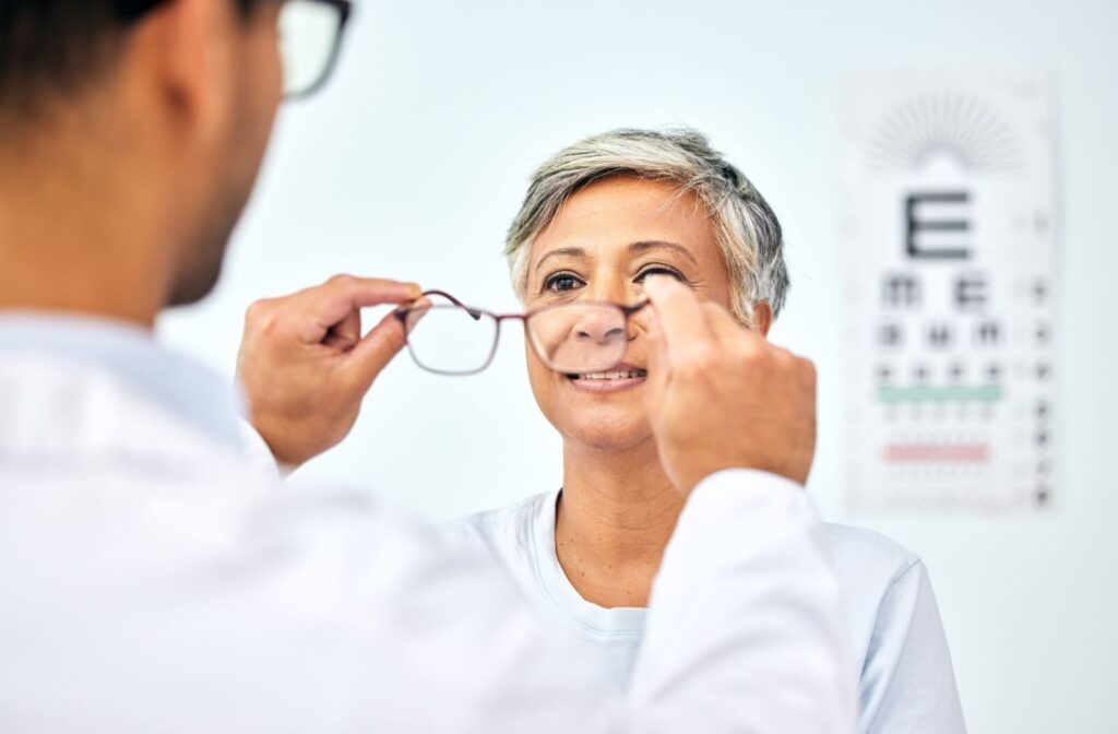 A smiling person with grey hair looking on as an optometrist holds up a pair of eyeglasses in front of their face.