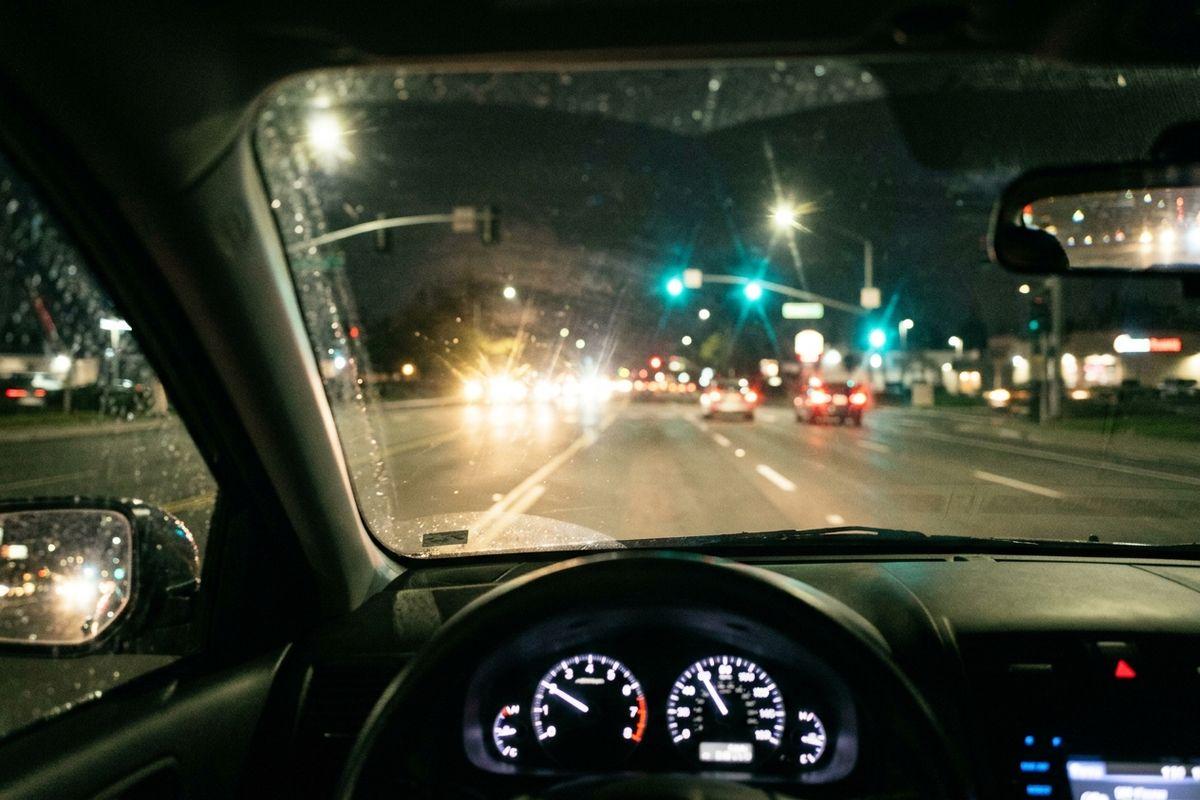 View from inside a car at night, dashboard and windshield visible, driving on a multi-lane road with street lights and green traffic signals ahead.